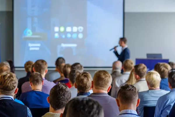 Audience at a presentation with speaker and screen.
